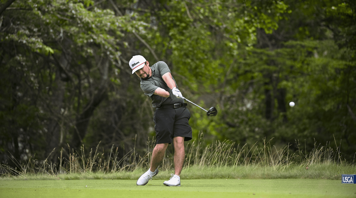 Golfer swinging club on lush course, ball in mid-flight amidst trees.