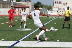 Soccer player in white jersey controlling ball on field during match, with referee and opponents in background.