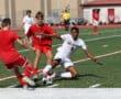 Soccer players in red and white uniforms vie for the ball on a sunny field during a competitive match.