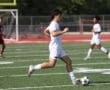 Soccer player in white kit dribbling on field during match, teammates and opponents in the background.