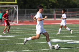 Soccer player in white kit dribbling on field during match, teammates and opponents in the background.
