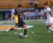 Soccer player in black and yellow Tigers uniform dribbles past white-clad defender on a green field during a match.