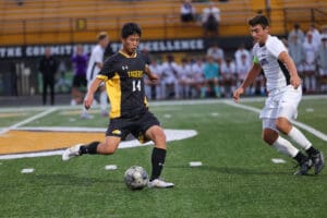 Soccer player in black and yellow Tigers uniform dribbles past white-clad defender on a green field during a match.
