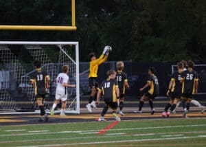 Soccer goalie catches a ball during a match, with players surrounding the goal post in a night game setting.