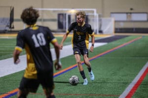 Soccer player in black and yellow uniform dribbles the ball on the field during a night match.