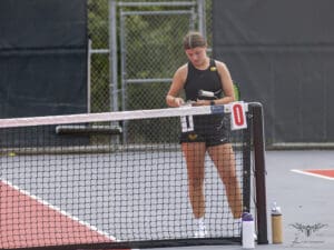 Tennis player prepares gear at the net of an outdoor court with scorecards and water bottles nearby.