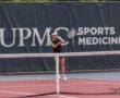 Tennis player hitting a forehand shot on an outdoor court with a sports medicine banner in the background.