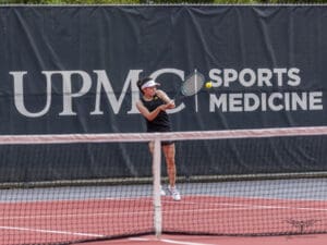 Tennis player hitting a forehand shot on an outdoor court with a sports medicine banner in the background.