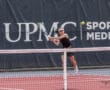 Young tennis player executing a backhand shot on a court, UPMC Sports Medicine banner in the background.