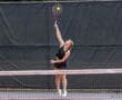 Tennis player in mid-air serve on court, wearing black outfit with focused expression.