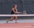 Tennis player hits a backhand shot during a match on a red court.
