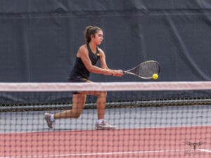 Tennis player hits a backhand shot during a match on a red court.