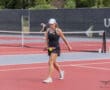 Young tennis player in action on a red court, wearing a black outfit and cap, holding a racket with a tennis ball.