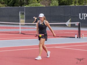 Young tennis player in action on a red court, wearing a black outfit and cap, holding a racket with a tennis ball.