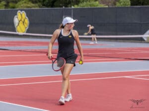Young tennis player in action on a red court holding a racket and ball, wearing black sportswear and a white cap.
