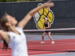 Tennis player prepares to return serve during a match on a bright red court, with a logo in the background.