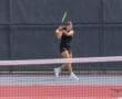 Tennis player in mid-swing during a match on a court with a net in the foreground.