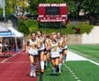 Girls' field hockey team in white uniforms jogging on the field with scoreboard in the background.