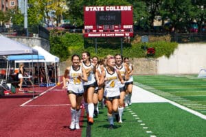 Girls' field hockey team in white uniforms jogging on the field with scoreboard in the background.