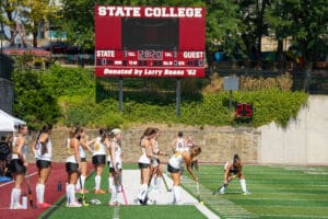 Field hockey team preparing for the match at State College, players in action near scoreboard, sunny day.