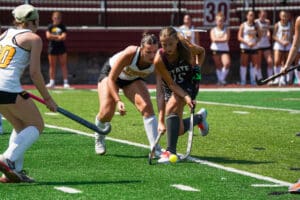 Two female field hockey players competing for the ball during a match on a sunny day.
