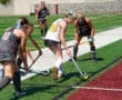 Women playing field hockey on a turf field, competing for the ball near a boundary line.