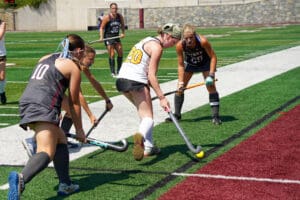 Women playing field hockey on a turf field, competing for the ball near a boundary line.