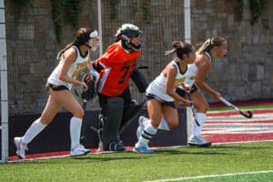 Field hockey players defend the goal during a match, wearing team uniforms and protective gear.
