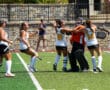 Field hockey team celebrates on turf after a victory. Players in white uniforms hug the goalie in orange gear.