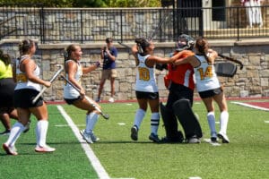 Field hockey team celebrates on turf after a victory. Players in white uniforms hug the goalie in orange gear.