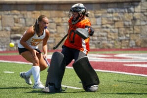 Field hockey goalkeeper blocking a shot as an opposing player approaches on a sunny day.