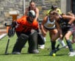 Field hockey players compete intensely near goal, with a goalkeeper in orange ready to defend.