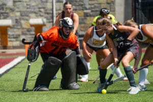 Field hockey players compete intensely near goal, with a goalkeeper in orange ready to defend.