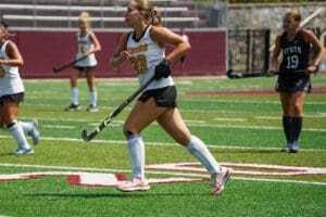 Field hockey players in action on a green turf, wearing team uniforms and protective gear.