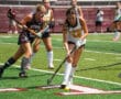 Field hockey game in action, female players competing on turf, player in white jersey dribbling ball.