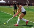Field hockey player in action, striking the ball during a competitive match on a sunny day.