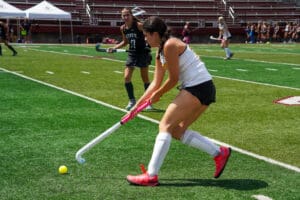 Field hockey player in action, striking the ball during a competitive match on a sunny day.