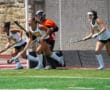 Field hockey players in action on a sunny day, wearing white and blue uniforms, near the goal area.