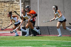 Field hockey players in action on a sunny day, wearing white and blue uniforms, near the goal area.