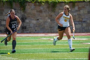 Two field hockey players competing intensely on a green field, one wearing State jersey, the other Tigers.