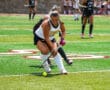 Field hockey players compete for the ball on a sunny sports field.