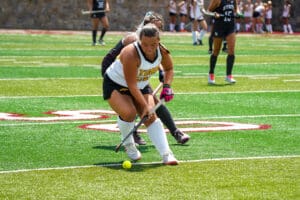 Field hockey players compete for the ball on a sunny sports field.