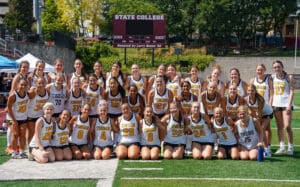 Girls' field hockey team posing on the field, wearing Tigers jerseys, in front of a scoreboard at State College.