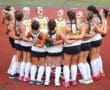 Girls' field hockey team in a huddle, wearing white jerseys, bonding before a game on a track field.