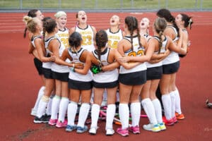 Girls' field hockey team in a huddle, wearing white jerseys, bonding before a game on a track field.