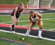 Two field hockey players compete for the ball during a match on a green turf field.