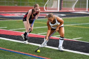 Two field hockey players compete for the ball during a match on a green turf field.