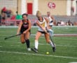 Two field hockey players compete for the ball on a green turf field during a match.