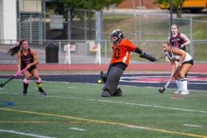 Girls' field hockey match with goalie defending and players in action on the field.
