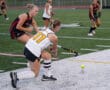 Field hockey players in action, player in white and black uniform about to hit the ball on a green turf field.